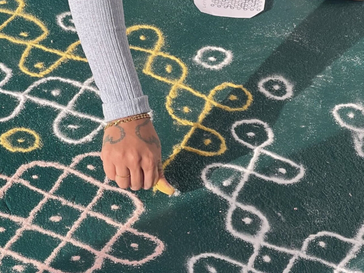 A brown tattooed hand with gold jewelry reaches down to draw a kolam on teal floor. The index finger holds rice flour or turmeric, trailing powder to form curving loops. Completed white and golden loops already surround the hand.